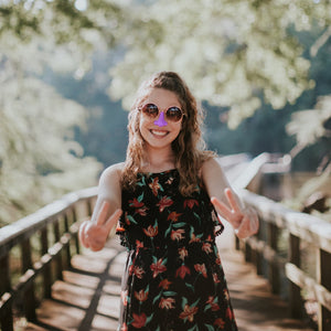 woman on a bridge in a forest wearing Nöz purple sunscreen on her nose holding out both arms giving peace signs